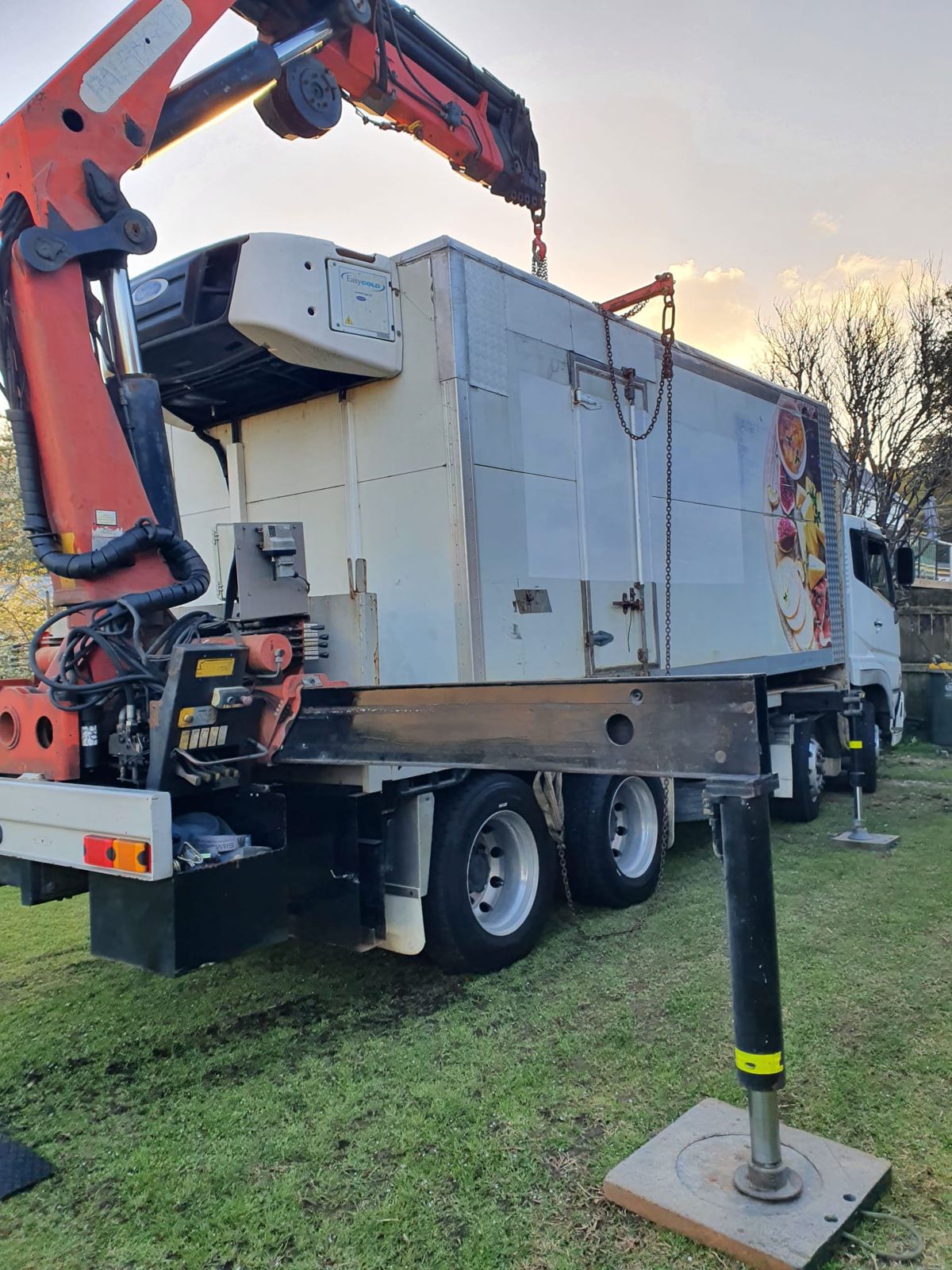 Rear of the crane truck on a lawn, outrigger leg down on a pad, the crane lifting a refrigerated truck body